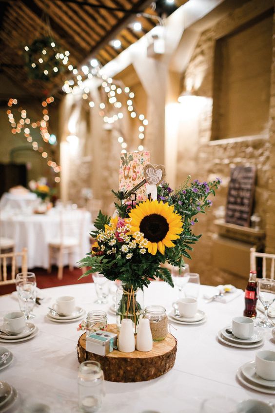 a lovely wedding centerpiece with wildflowers, fern and a sunflower, thistles, a burlap heart placed on a wood slice is a gorgeous idea
