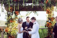 a lush fall rustic wedding arch with lots of greenery, orange, red blooms and berries and lots of twigs and cascading details is amazing