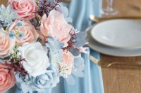 a pretty wedding tablescape with a blue table runner, pink, blue and white blooms and candles plus matching gold cutlery