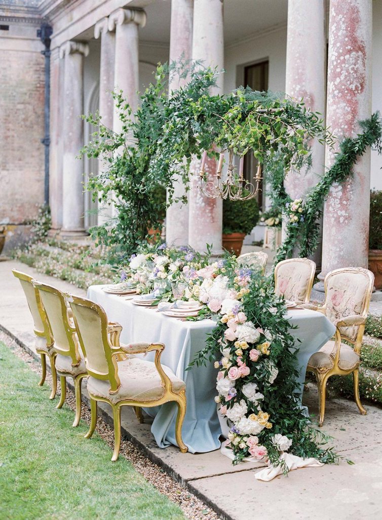 A refined garden wedding tablescape with a lush greenery and pastel bloom runner, pastel plates and linens and refined chairs