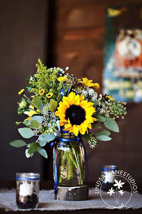 a relaxed rustic wedding centerpiece of eucalyptus, sunflowers, white and yellow blooms and candles around is a beautiful summer solution
