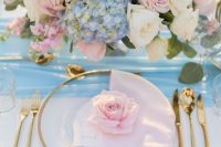 a romantic wedding tablescape with a blue table runner, pink napkins, pink and white roses, blue hydrangeas, greenery and gold cutlery