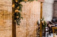 a rust fall wedding arch with greenery and peachy blooms, candle lanterns and a light curtain behind it is a very chic and cool idea