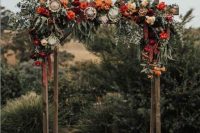 a rustic fall wedding arch decorated with greenery, orange, red, burgundy and pink blooms and much texture and dimension