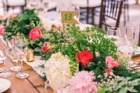 a stylish secret garden wedding tablescape with a greenery and pink bloom runner, gold candleholders, glasses and a white table runner