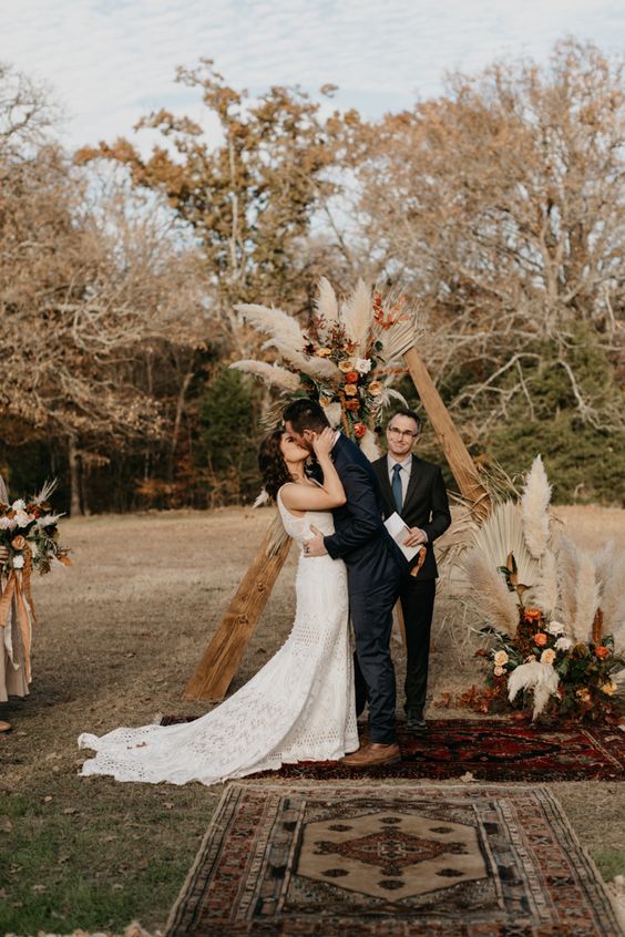 a triangle boho chic fall wedding arch with dried fronds, pampas grass, blush, orange and rust-colored blooms and boho rugs