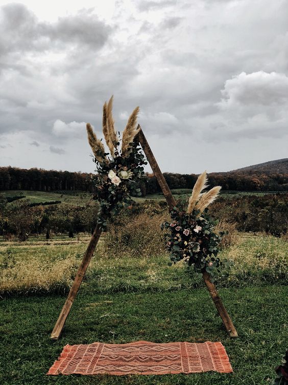 a triangle boho fall wedding arch with greenery, white and blush blooms, pampas grass and a boho rug is cool