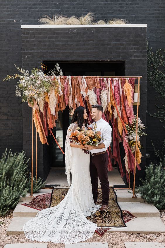 a very creative boho fall wedding arch done with long burgundy, orange, rust and pink ribbons, some white and yellow blooms and greenery