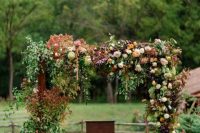 a very lush rustic fall wedding arch decorated with greenery and dried foliage, with blush, rust, deep purple, white blooms and pampas grass