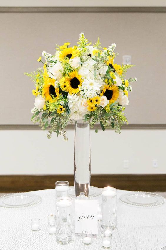an elegant colorful wedding centerpiece of greenery, billy balls, sunflowers, white roses and other blooms in a tall glass vase