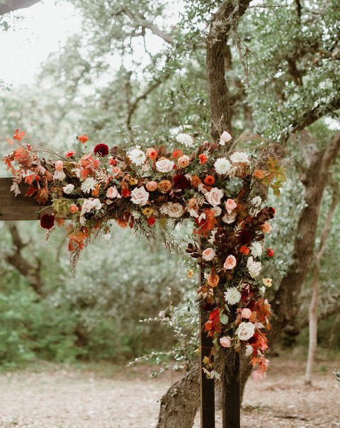 an elegant rustic fall wedding arch detailed with blush, red, rust, orange, burgundy blooms, greenery and bright fall foliage