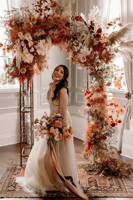 an incredible bright boho fall wedding arch with rust-colored and pink and blush roses, white orchids, pampas grass, rust-colored fall foliage just excites