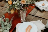a beautiful boho fall wedding centerpiece of orange, burgundy, white blooms, greenery, a rust-colored table runner and candles around