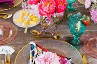 a bright bridal shower tablescape with floral print napkins, bold blooms in hot pink and pink, gold cutlery, blue glasses and pink glasses is cool