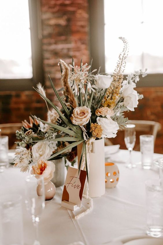 a catchy fall boho wedding centerpiece of a white vase, greenery, blush, rust and white blooms, pampas grass and candles around