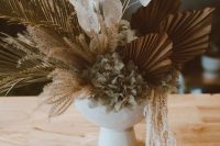 a chic wedding centerpiece of leaves, fronds, lunaria, dried hydrangeas in a white bowl is a gorgeous idea for a boho wedding