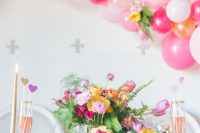 a colorful bridal shower tablescape with yellow and hot pink blooms and greenery, pink chargers, a colorful balloon garland and gold candles, floral pillows