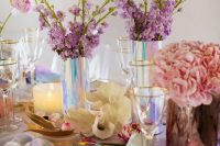 a holographit wedding table setting with a white sequin tablecloth, a clear charger, lilac and pink blooms and gold cutlery