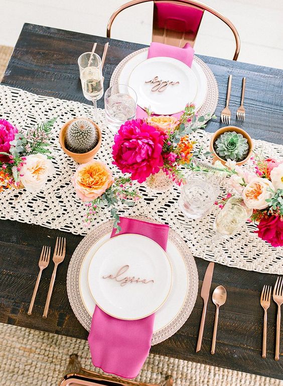 a lovely bold bridal shower tablescape with a macrame runner, pink napkins and blooms, cacti and succulents and copper cutlery