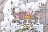 a refined vintage sweets table of a white dresser, with greenery, white blooms and a white balloon garland with greenery is gorgeous