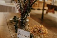 a simple and relaxed fall boho wedding centerpiece of a glass jar with dried flasses, leaves, greenery on the table and dried blooms, a macrame runner