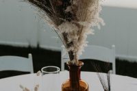 a simple boho fall wedding centerpiece of an apothecary bottle with dried grasses and feathers, candles around is a veyr easy to recreate idea