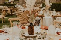 a summer or fall wedding centerpiece of an apothecary bottle, dried fronds, leaves and a wood slice plus greenery is a lovely idea