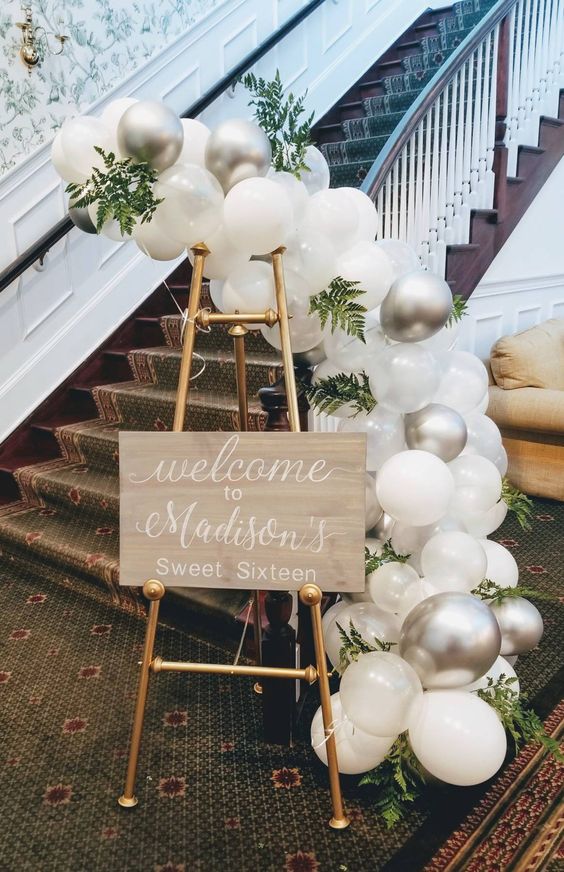 a welcome sign on gold legs, with clear, white and silver balloons and greenery is a very pretty and chic idea to rock