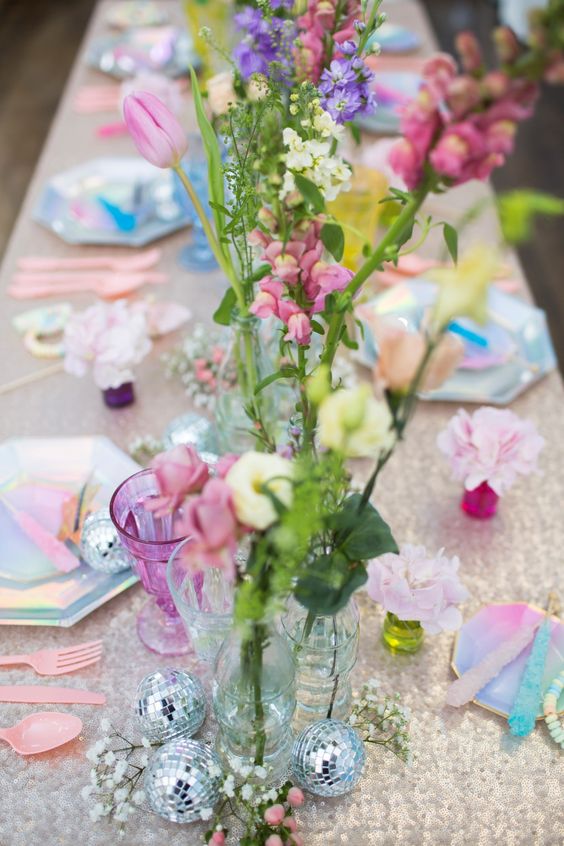 an iridescent wedding tablescape with a white sequin tablecloth, pink, lilac and white blooms, disco balls, iridescent plates and sugar crystal candies