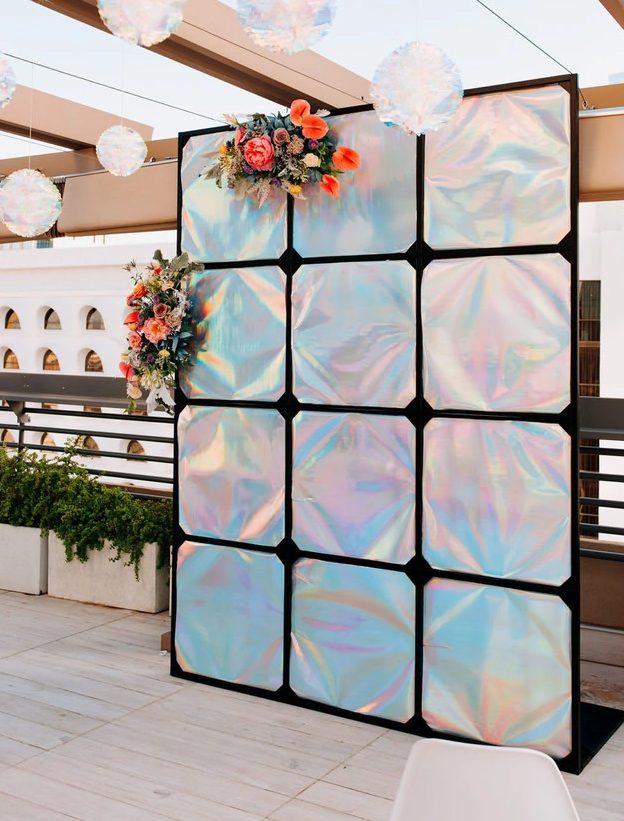 an iridescent holographic wedding backdrop decorated with pink, orange blooms and greenery is a stylish solution for an iridescent wedding