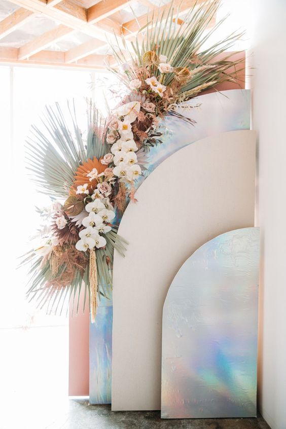 an iridescent wedding backdrop with rounded holographic and blush screens and iridescent wedding florals and fronds