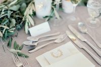 a beautiful greige wedding table setting with a greige tablecloth, white linens and cards, a greenery garland and pillar candles is chic