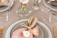 a chic and delicate wedding tablescape done with greige linens and plates, with pink napkins and blooms and gold cutlery is chic