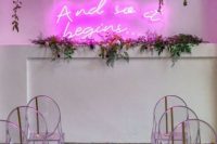 a chic bright minimalist wedding ceremony space with a pink neon sign, greenery, hanging blooms and acrylic chairs