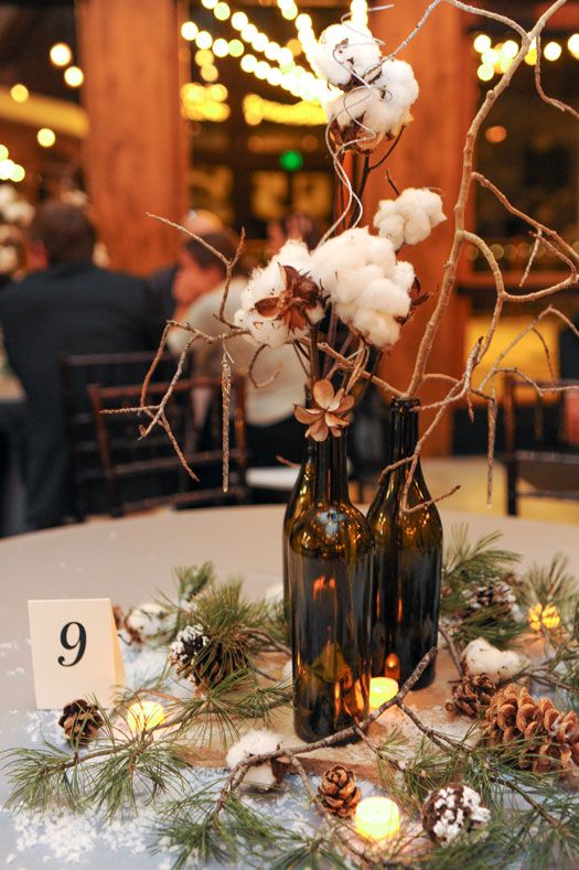 a cozy winter centerpiece with snowy pinecones, evergreens, candles, cottons and wine bottles with branches and faux snow around it