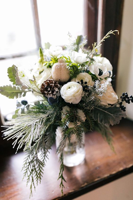 a dreamy winter wedding bouquet of evergreens, pale leaves, white anemones and ranunculus, privet berries, thistles and snowy pinecones