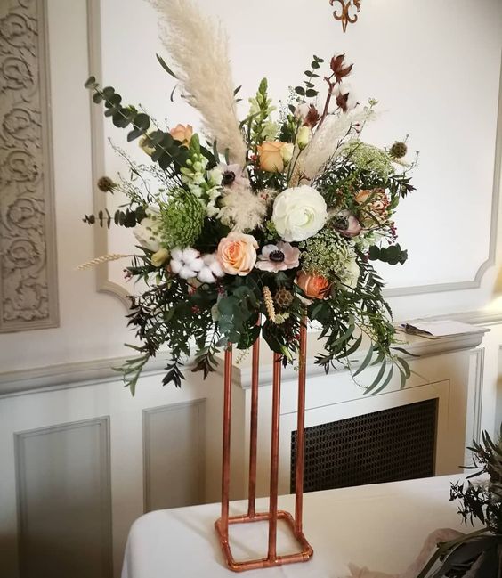 a gorgeous tall wedding centerpiece of white and peachy blooms, pampas grass, lots of greenery, seed pods, cotton and foliage on a copper stand