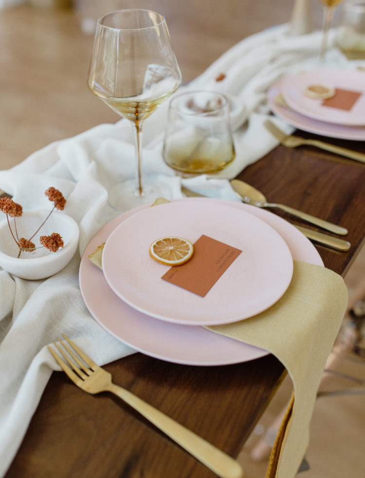 a minimalist bright wedding tablescape with a neutral runner, pink plates, mustard napkins and gold cutlery, amber glasses