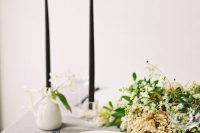 a pretty and elegant minimalist wedding tablescape with a grey tablecloth and white napkins, a marble charger, black candles and a greenery and white bloom centerpiece