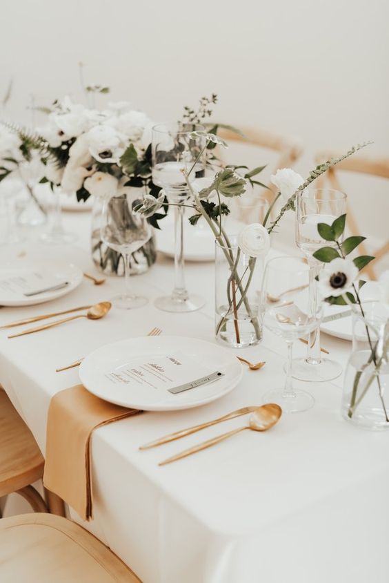 a pretty scandinavian wedding tablescape with white blooms and greenery, white plates, gold cutlery and amber napkins plus floating candles