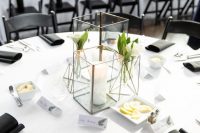 a stylish minimalist wedding tablescape with candle lanterns with blooms and a candle, with black napkins and a white tablecloth