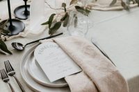a stylish neutral wedding tablescape with a minimalist feel, with neutral plates and textural linens, greenery, blue candles and acrylic table numbers