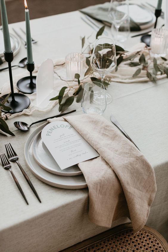 a stylish neutral wedding tablescape with a minimalist feel, with neutral plates and textural linens, greenery, blue candles and acrylic table numbers