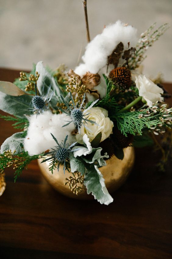 a textural and cool wedding centerpiece of a gilded vase with greenery, thistles, cotton, white and dried dark blooms and foliage is gorgeous