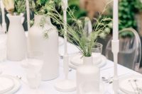 a very simple and minimal wedding tablescape with all white everything, with greenery and white blooms, tall and thin white candles and some cutlery