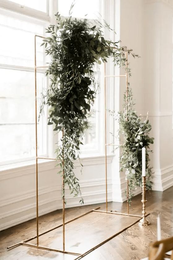 a brass minimalist wedding arch decorated with lush greenery and white blooms plus a single candle in a tall candlestick is awesome