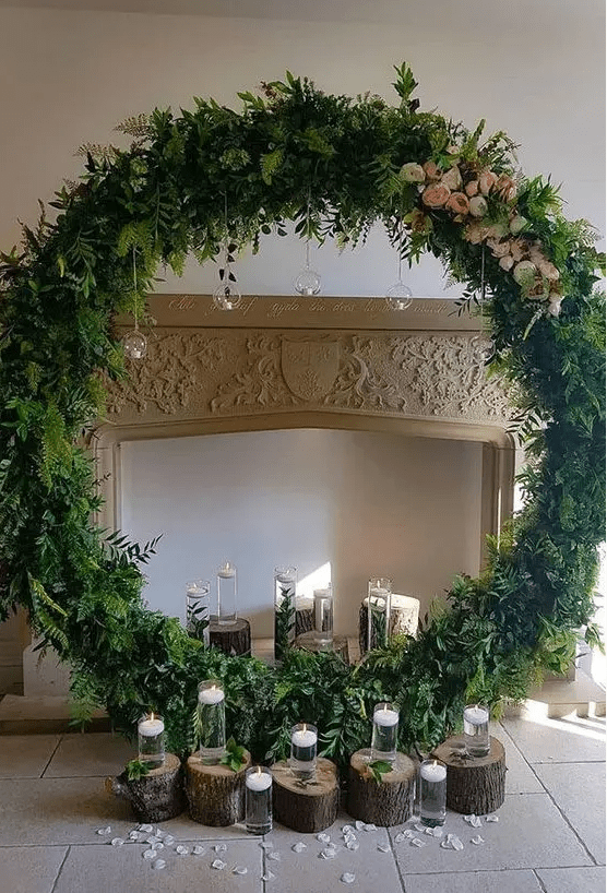 a faux fireplace, a greenery round altar with blooms and candles on stumps and hanging down from the wreath