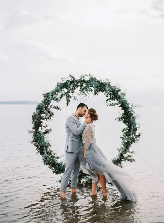 a greenery round wedding arch placed right in the sea is a unique and creative idea for a coastal wedding