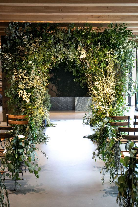 a jaw-dropping greenery wedding arch covered with various types of greenery and branches and matching greenery on the aisle chairs