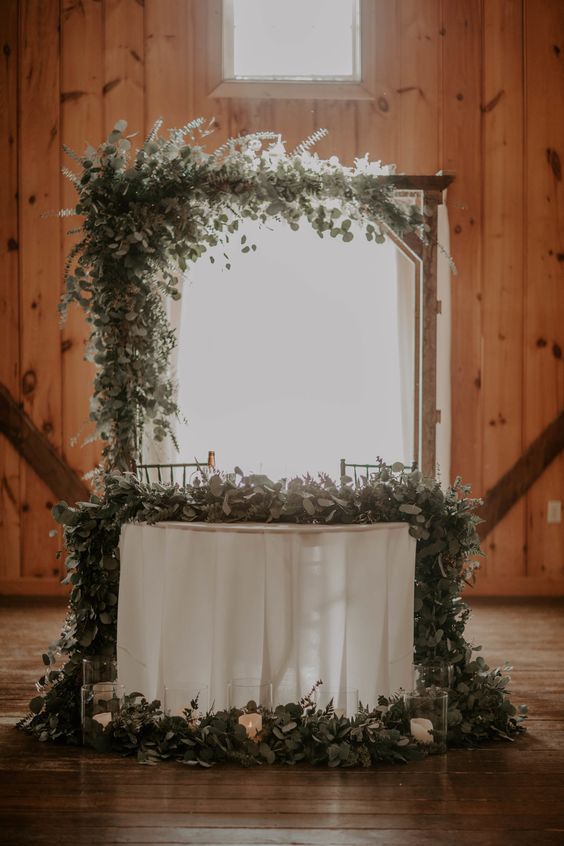 a lovely wedding arch of wood covered with various types of eucalyptus and fern and a matching garland on the table and around it